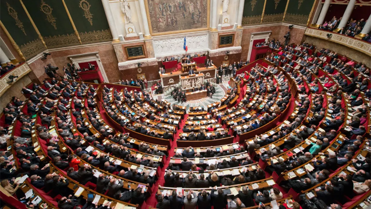 French National Assembly chamber during a budget vote, with streaming icons representing a per-gigabyte tax on tech platforms.