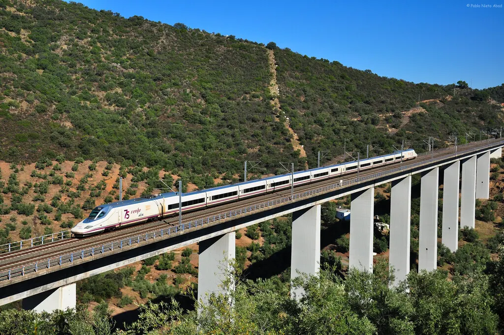 A high-speed train travelling along a Spanish railway line with trackside signalling equipment.