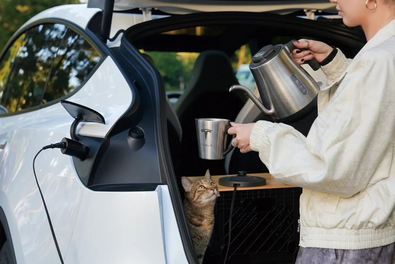 A Tesla electric car parked at a campsite, powering an electric kettle through a plug-in adapter connected to its charge port.