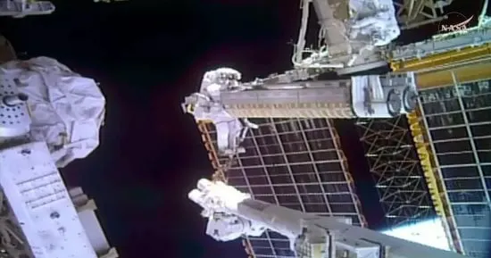 Two astronauts in spacesuits install a folding iROSA solar array on the exterior of the International Space Station during a spacewalk.