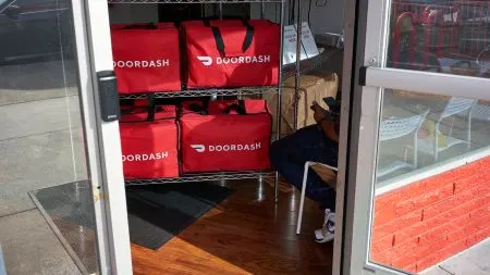 A delivery courier carrying grocery bags and a food order to a customer's front door.